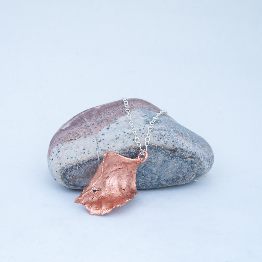 Copper leaf resting on one of its curved sides, leaning on grey side of grey, white and red beach stone