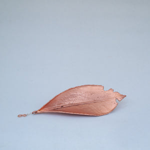 Large copper leaf, laid on its back, on flat surface, with silver chain and copper 8 poking out near the stem