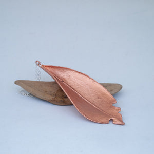 Copper laurel leaf resting on a piece of driftwood, silver chain behind, white background