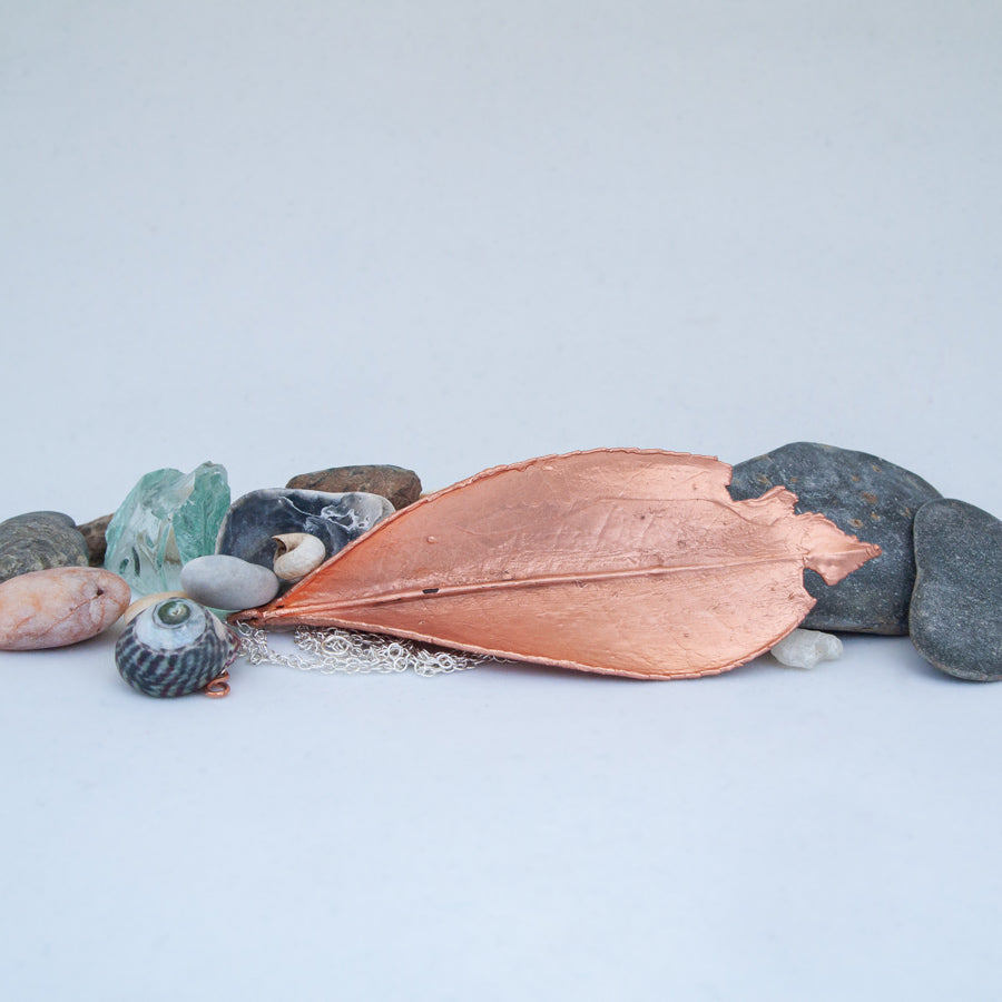 Laurel leaf in copper, laid on side surrounded by beach pebbles, glass and shells