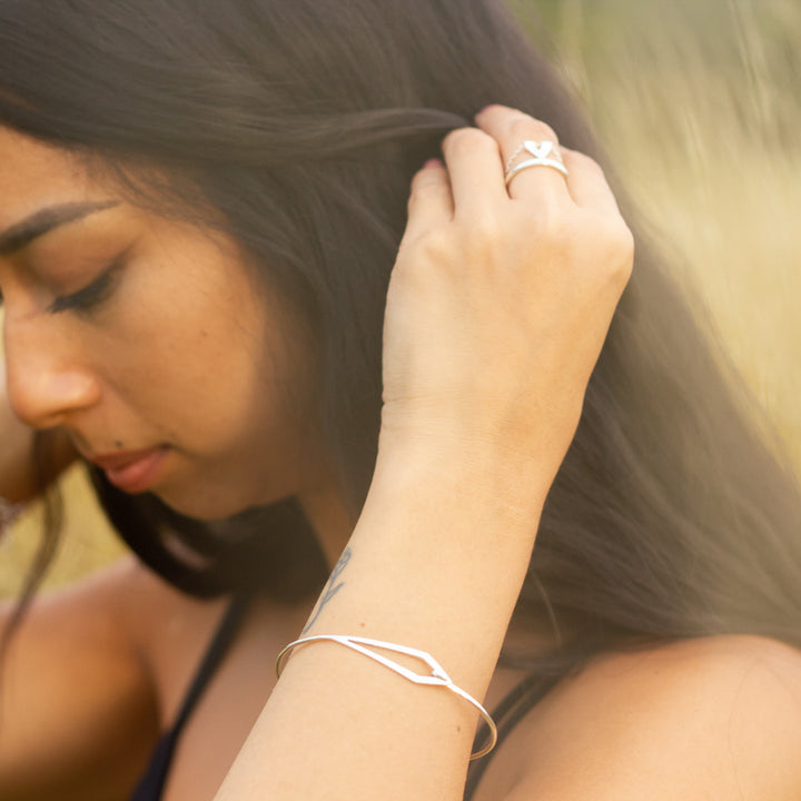 Unusual silver handmade bangle on woman's arm, as she pulls back her dark hair to her ears, in a field of yellow grass