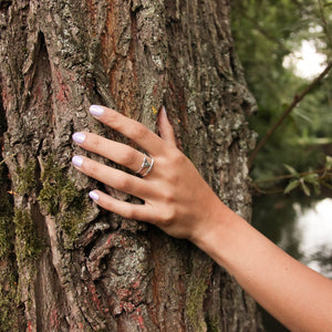 Tailwind ring worn on third finger of model's left hand against the brown green red bark of a tree next to the river