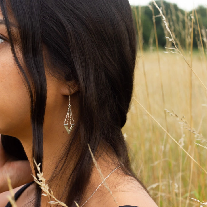 Unusual handmade silver earrings of Zephyr design dangling from ear lobe of model with hair swept behind shoulder in field of yellow grass